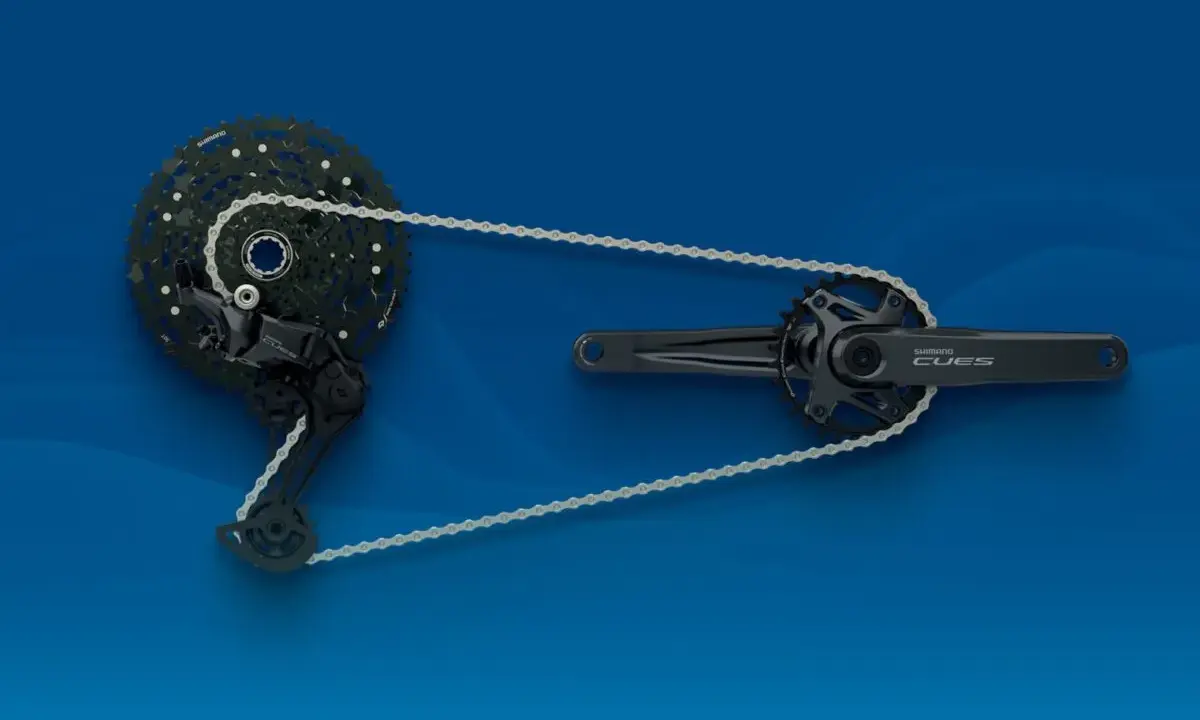 Zestaw napędowy **Shimano CUES**: przerzutka tylna, kaseta i korba z zębatkami, połączone łańcuchem na niebieskim tle.