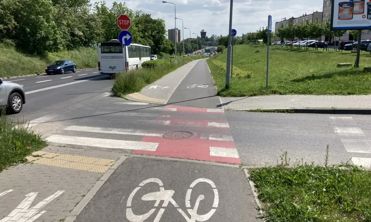Pasy na ścieżce rowerowej i pasy dla pieszych przecinają drogę. Autobus i samochody czekają na zielone światło.