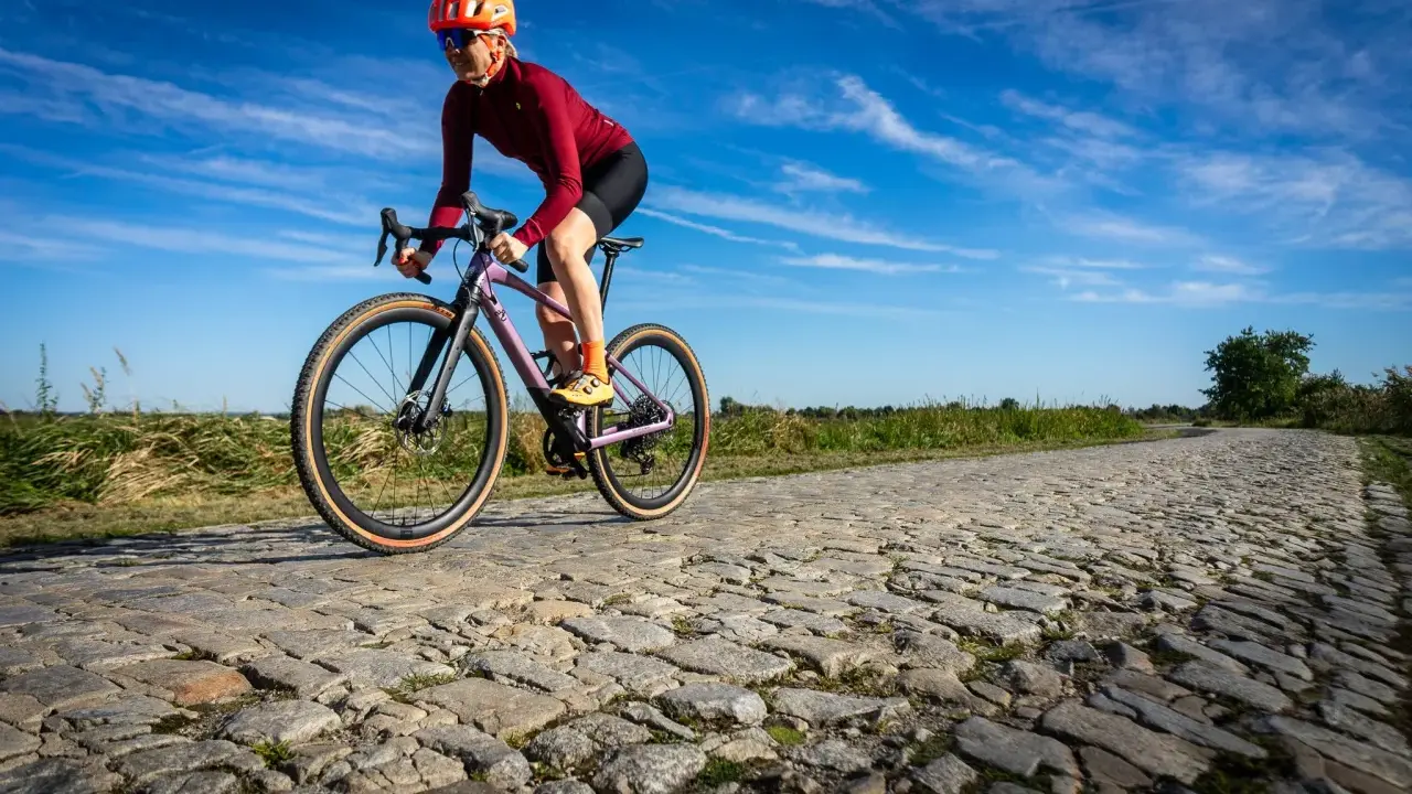 Kobieta w kasku i okularach jedzie na rowerze BMC URS LT po brukowanej drodze.