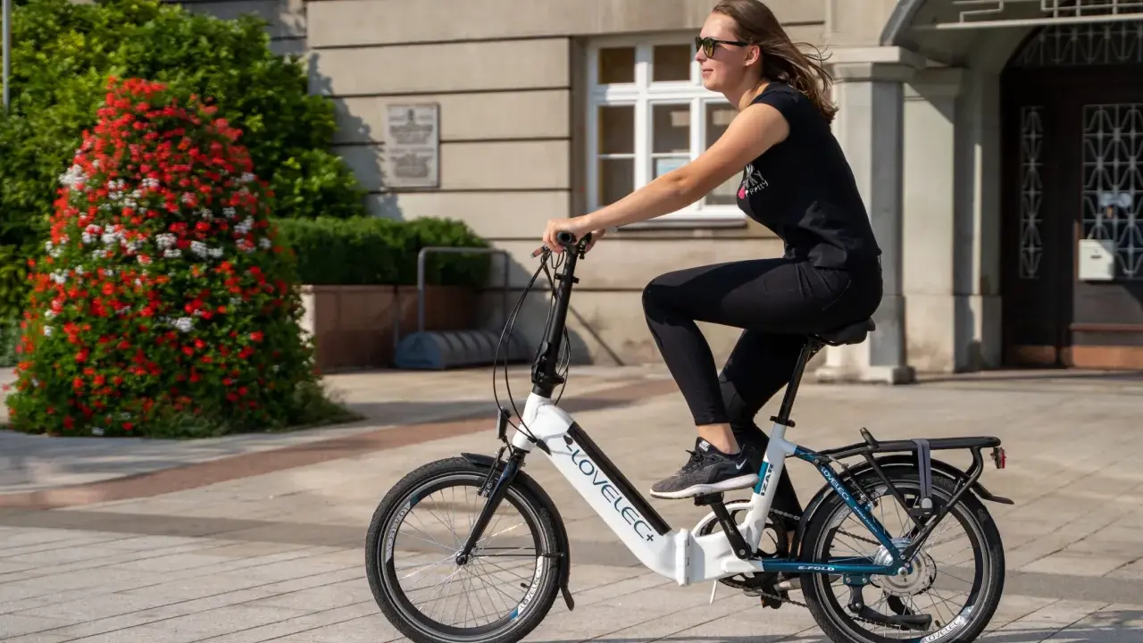 Młoda kobieta w okularach przeciwsłonecznych i czarnej koszulce jedzie na rowerze elektrycznym LOVELEC+. To jazda rowerem elektrycznym bez wspomagania, po brukowanym chodniku, obok krzewu z czerwonymi kwiatami.