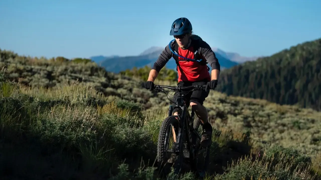 Kolarz MTB w kasku i czerwonej koszulce z plecakiem wodnym, jadący po górskim szlaku. Utrzymuje prawidłową pozycję na rowerze mtb, pochylony do przodu.