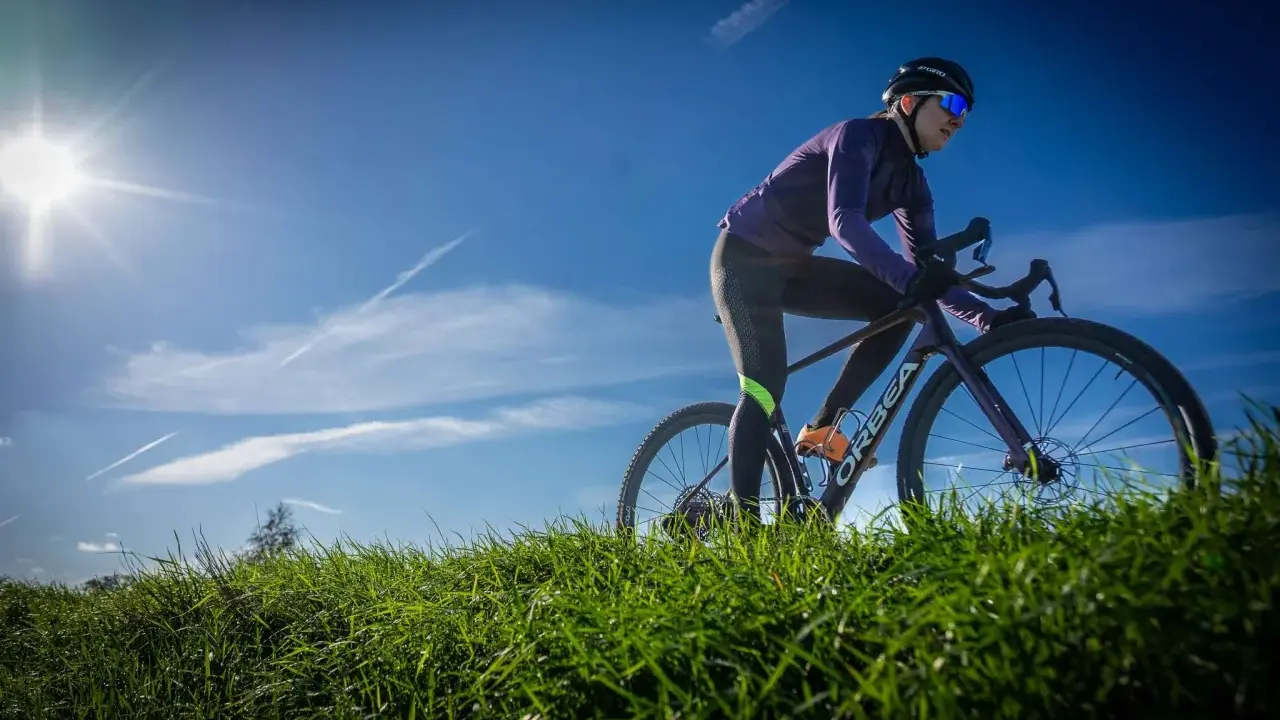 Kobieta w kasku i okularach przeciwsłonecznych odbywa trening na rowerze gravelowym, pokonując pagórkowaty teren porośnięty zieloną trawą.