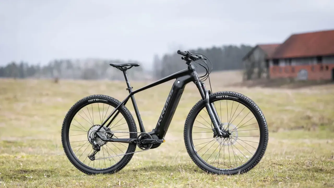 Czarny rower Kross Level Boost 3.0 z napędem elektrycznym stoi na trawie. W tle budynki i drzewa.