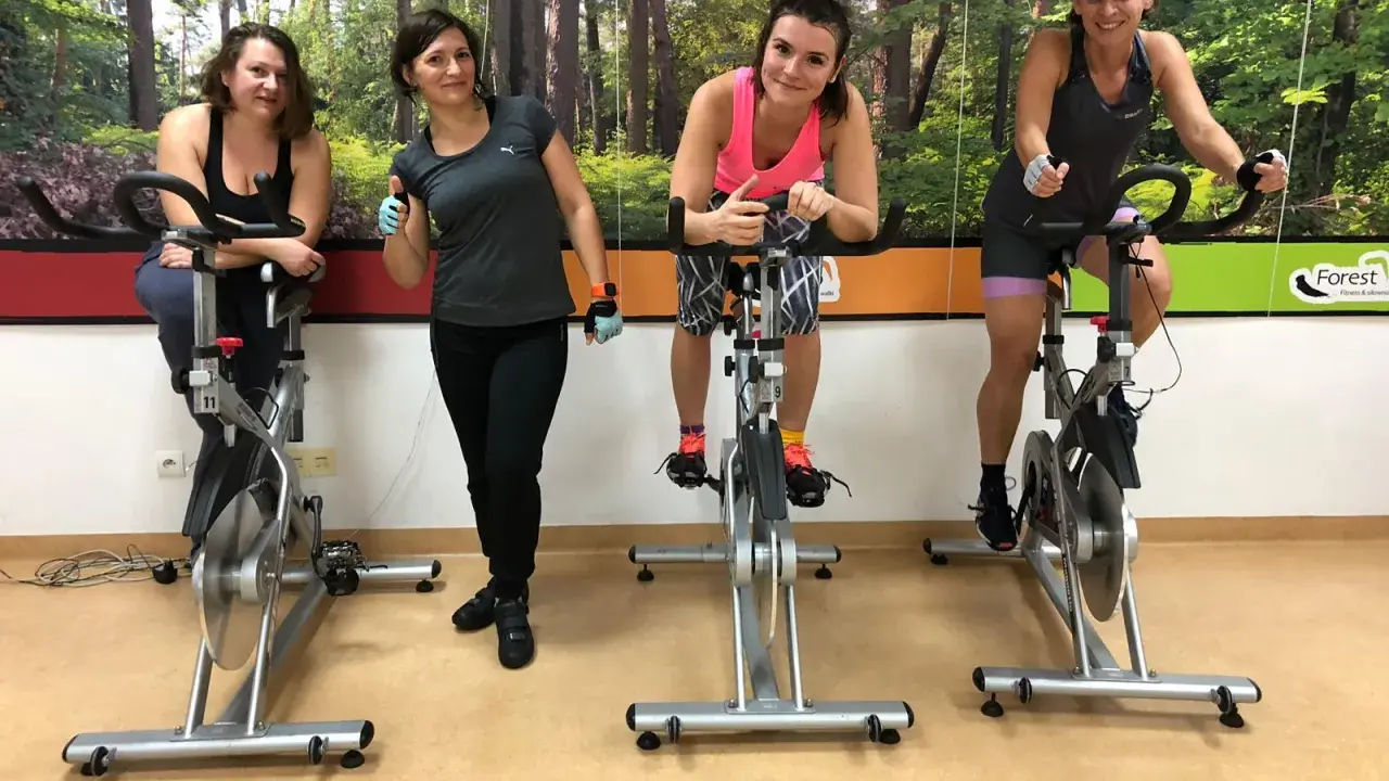 Cztery kobiety ćwiczą na rowerach stacjonarnych. W tle las. To świetny sposób na trening spinning co to jest?