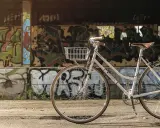 Biały rower z koszykiem, idealny jako rower miejski czy trekkingowy, stoi na tle ściany pokrytej kolorowym graffiti.