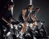 Trzy osoby ćwiczą na rowerach stacjonarnych w ciemnym pomieszczeniu. Widać ich skupienie i wysiłek, co sugeruje pozytywne indoor cycling efekty.