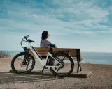 Kobieta odpoczywa obok białego roweru elektrycznego, który jest jednym z wielu rodzajów rowerów elektrycznych. Widok na morze.