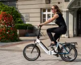 Młoda kobieta w okularach przeciwsłonecznych i czarnej koszulce jedzie na rowerze elektrycznym LOVELEC+. To jazda rowerem elektrycznym bez wspomagania, po brukowanym chodniku, obok krzewu z czerwonymi kwiatami.