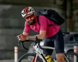 Mężczyzna w różowej koszulce i czarnych spodenkach, kasku i okularach rowerowych, gotowy na jazdę przy 10 stopniach. Plecak na plecach.