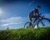 Kobieta w kasku i okularach przeciwsłonecznych odbywa trening na rowerze gravelowym, pokonując pagórkowaty teren porośnięty zieloną trawą.
