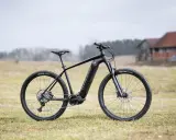 Czarny rower Kross Level Boost 3.0 z napędem elektrycznym stoi na trawie. W tle budynki i drzewa.