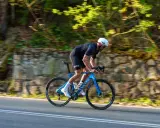 Kolarz w kasku ściga się na niebieskim rowerze Canyon Endurace CF 7 po malowniczej drodze wśród zieleni.