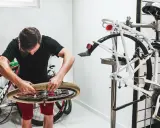 Mężczyzna w czarnym t-shircie i czerwonych spodenkach naprawia rower, przygotowując się do montażu chwytów na kierownicę.