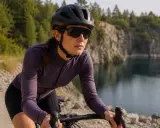 Kobieta w kasku i ciemnych okularach rowerowych jedzie rowerem szutrową drogą nad jeziorem. Idealne okulary na rower chronią oczy.