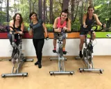 Cztery kobiety ćwiczą na rowerach stacjonarnych. W tle las. To świetny sposób na trening spinning co to jest?