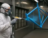 Pracownik w kombinezonie i masce maluje proszkowo niebieską ramę rowerową.