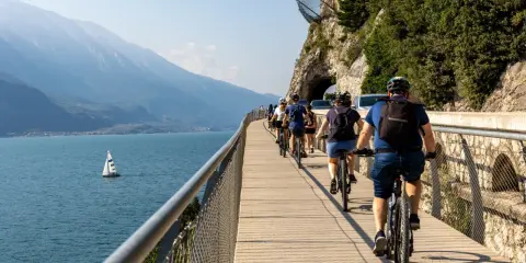Grupa rowerzystów przemierza malownicze trasy rowerowe wokół jeziora, podziwiając góry i żaglówkę.