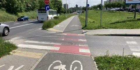 Pasy na ścieżce rowerowej i pasy dla pieszych przecinają drogę. Autobus i samochody czekają na zielone światło.