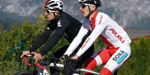 Dwóch kolarzy, być może najlepsi kolarze w historii, w biało-czerwonych strojach z napisem "Polska" i logiem "Bora Hansgrohe", ściga się na tle gór.