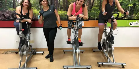 Cztery kobiety ćwiczą na rowerach stacjonarnych. W tle las. To świetny sposób na trening spinning co to jest?