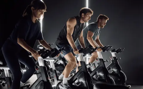 Trzy osoby ćwiczą na rowerach stacjonarnych w ciemnym pomieszczeniu. Widać ich skupienie i wysiłek, co sugeruje pozytywne indoor cycling efekty.