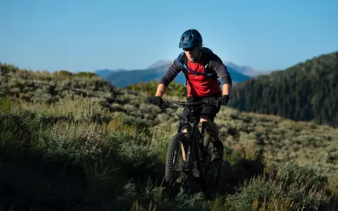 Kolarz MTB w kasku i czerwonej koszulce z plecakiem wodnym, jadący po górskim szlaku. Utrzymuje prawidłową pozycję na rowerze mtb, pochylony do przodu.