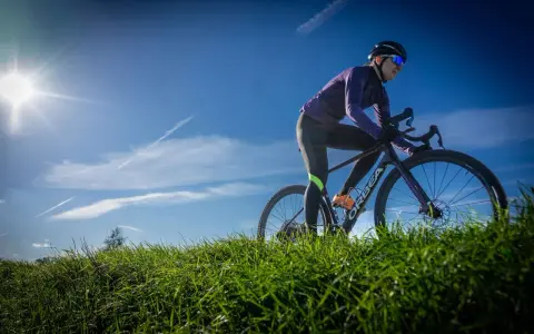 Kobieta w kasku i okularach przeciwsłonecznych odbywa trening na rowerze gravelowym, pokonując pagórkowaty teren porośnięty zieloną trawą.