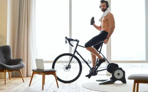 Mężczyzna w słuchawkach pije wodę, ćwicząc na rowerze stacjonarnym przy dobrej muzyce na rower.