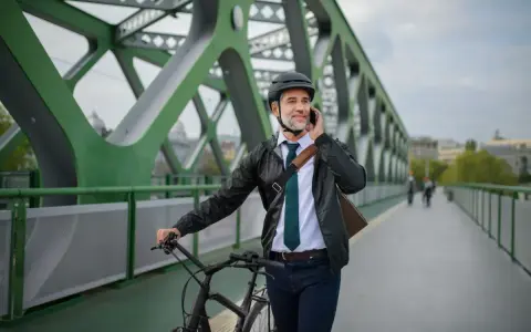 Mężczyzna w kasku i eleganckim stroju rozmawia przez telefon, prowadząc rower. Może to być jego nowy rower na firmę, idealny do miejskich dojazdów.