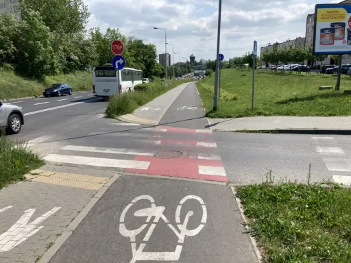 Pasy na ścieżce rowerowej i pasy dla pieszych przecinają drogę. Autobus i samochody czekają na zielone światło.