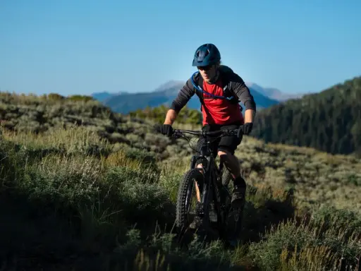 Kolarz MTB w kasku i czerwonej koszulce z plecakiem wodnym, jadący po górskim szlaku. Utrzymuje prawidłową pozycję na rowerze mtb, pochylony do przodu.