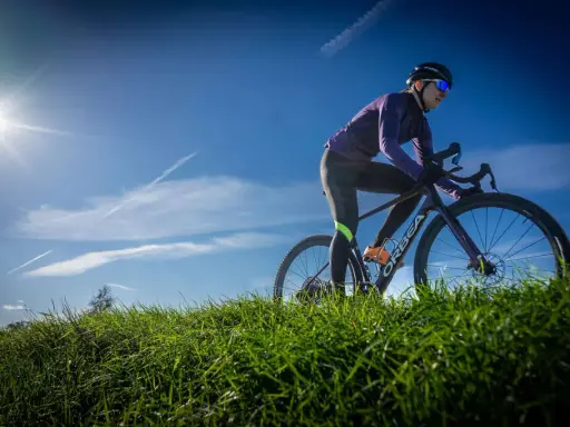 Kobieta w kasku i okularach przeciwsłonecznych odbywa trening na rowerze gravelowym, pokonując pagórkowaty teren porośnięty zieloną trawą.