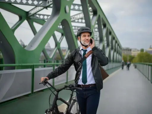 Mężczyzna w kasku i eleganckim stroju rozmawia przez telefon, prowadząc rower. Może to być jego nowy rower na firmę, idealny do miejskich dojazdów.