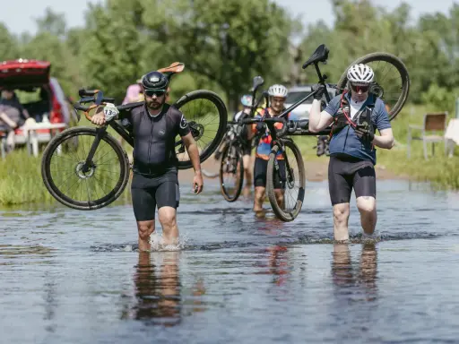 Kolarze pokonują wodną przeszkodę podczas mazowiecki gravel. Niosą rowery przez płytką rzekę, w tle zielone drzewa i samochód.
