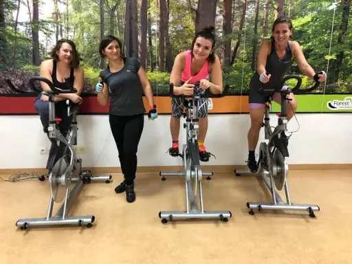 Cztery kobiety ćwiczą na rowerach stacjonarnych. W tle las. To świetny sposób na trening spinning co to jest?