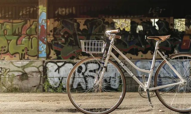Biały rower z koszykiem, idealny jako rower miejski czy trekkingowy, stoi na tle ściany pokrytej kolorowym graffiti.