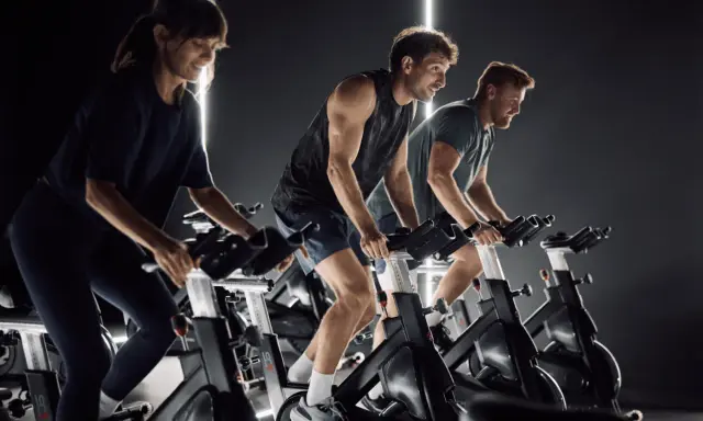 Trzy osoby ćwiczą na rowerach stacjonarnych w ciemnym pomieszczeniu. Widać ich skupienie i wysiłek, co sugeruje pozytywne indoor cycling efekty.