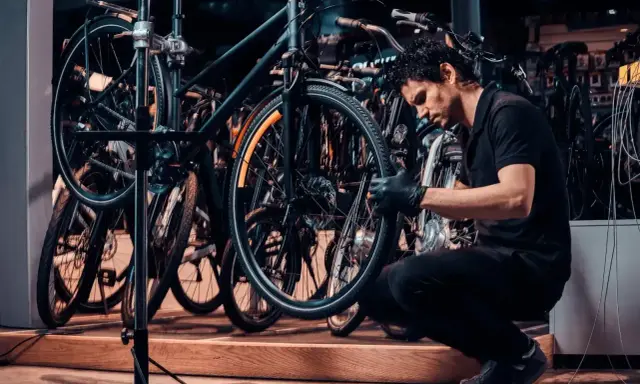 Mechanik w czarnej koszulce i rękawiczkach pokazuje, jak zdjąć tylne koło w rowerze, pracując w serwisie rowerowym.