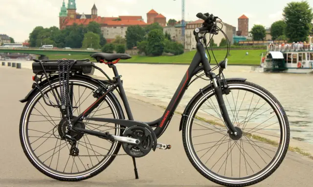 Czarny rower elektryczny z baterią na bagażniku, gotowy do jazdy po bulwarach z widokiem na Wawel.