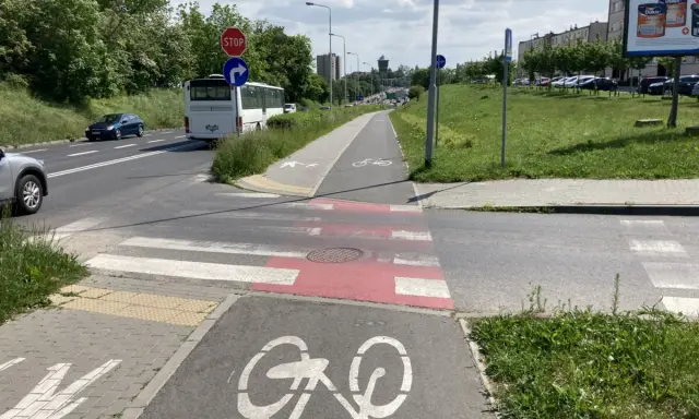 Pasy na ścieżce rowerowej i pasy dla pieszych przecinają drogę. Autobus i samochody czekają na zielone światło.