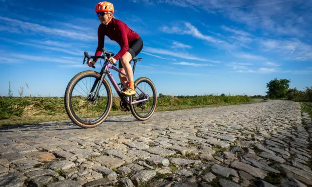 Kobieta w kasku i okularach jedzie na rowerze BMC URS LT po brukowanej drodze.