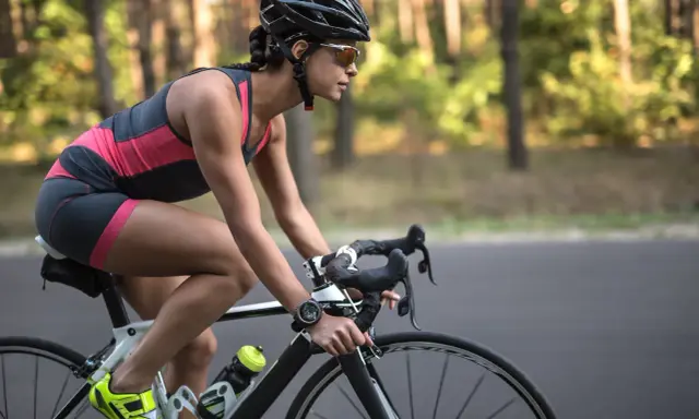 Kobieta w kasku i okularach przeciwsłonecznych pedałuje na rowerze szosowym przez las. Widać jej nogi w sportowym stroju i zegarek na ręce. To efekty po miesiącu regularnej jazdy na rowerze.