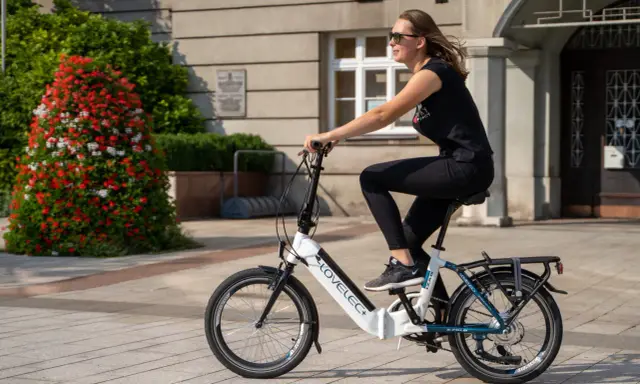 Młoda kobieta w okularach przeciwsłonecznych i czarnej koszulce jedzie na rowerze elektrycznym LOVELEC+. To jazda rowerem elektrycznym bez wspomagania, po brukowanym chodniku, obok krzewu z czerwonymi kwiatami.