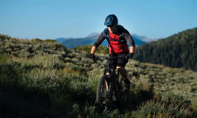 Kolarz MTB w kasku i czerwonej koszulce z plecakiem wodnym, jadący po górskim szlaku. Utrzymuje prawidłową pozycję na rowerze mtb, pochylony do przodu.