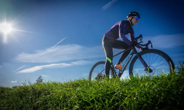 Kobieta w kasku i okularach przeciwsłonecznych odbywa trening na rowerze gravelowym, pokonując pagórkowaty teren porośnięty zieloną trawą.
