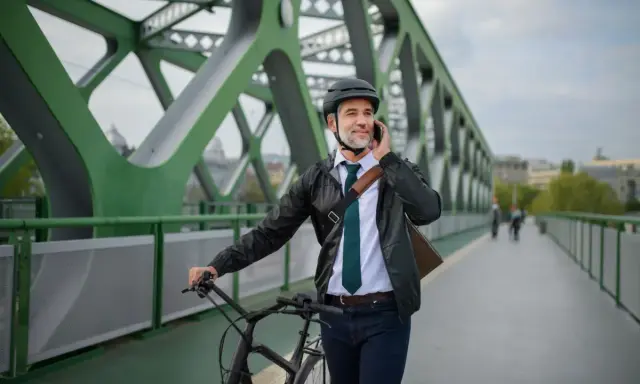 Mężczyzna w kasku i eleganckim stroju rozmawia przez telefon, prowadząc rower. Może to być jego nowy rower na firmę, idealny do miejskich dojazdów.