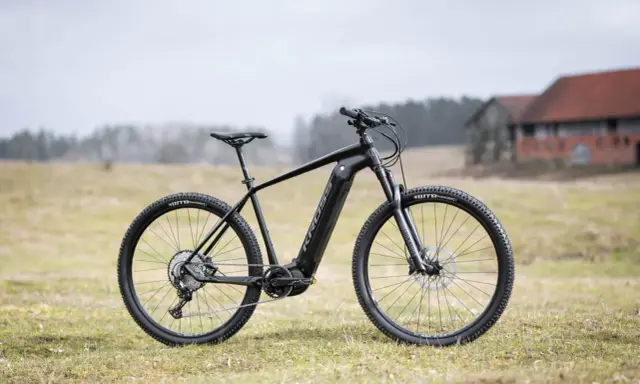Czarny rower Kross Level Boost 3.0 z napędem elektrycznym stoi na trawie. W tle budynki i drzewa.
