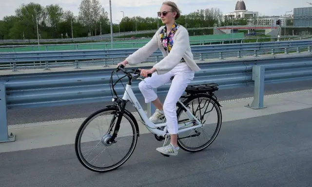 Kobieta na rowerze elektrycznym, ubrana na biało, jedzie po moście. Uważaj na przepisy, by nie dostać mandatu za rower elektryczny.