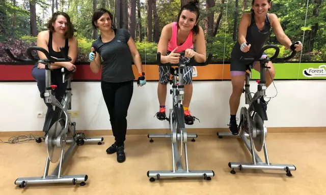 Cztery kobiety ćwiczą na rowerach stacjonarnych. W tle las. To świetny sposób na trening spinning co to jest?