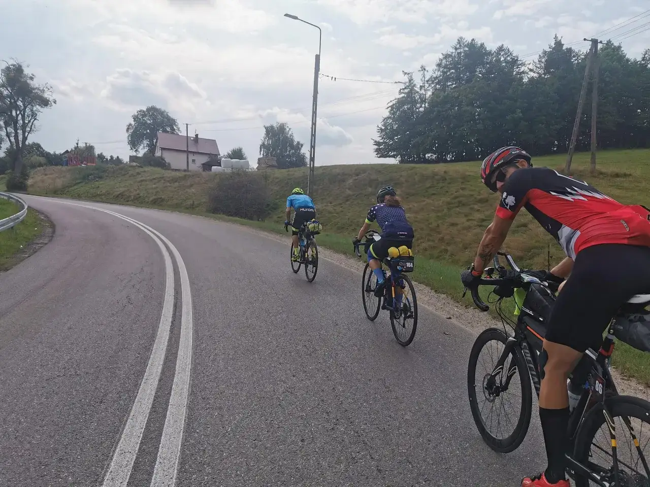 Trzech kolarzy pokonuje malowniczą trasę podczas maratonu rowerowego. Słońce przebija się przez chmury, a droga wije się wśr&oacute;d zielonych wzg&oacute;rz.